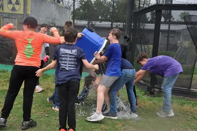 A group of enthusiastic participants engage in a spirited ice bucket challenge during an outdoor event, creating moments of laughter and camaraderie as water splashes across the grass. The colorful mix of team jerseys and casual attire captures the fun, competitive spirit of the gathering in what appears to be a recreational sports or charity event.