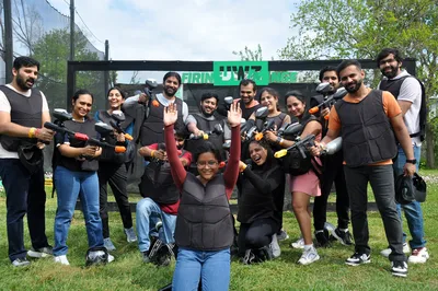 A large group of paintball enthusiasts gathers outdoors with their gear and weapons, smiling and posing together on a grassy field beneath overcast skies. The diverse team displays camaraderie and excitement, captured in a moment of celebration after what appears to be an intense recreational paintball competition.